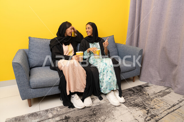 Enjoying free time, a group home entertainment activity, having fun eating popcorn while watching TV, spending quality time with friends, two Emirati Gulf Arab women wearing abaya and hijab sitting on the couch watching an entertaining movie with gestures of integration and pleasure