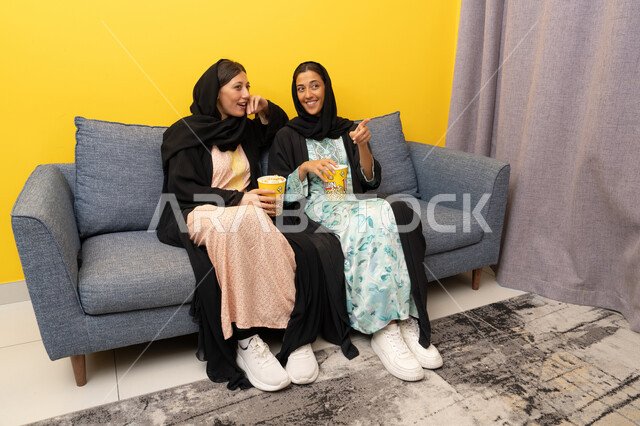 Group home entertainment activity, having fun eating popcorn while watching TV, spending quality time with friends, two Emirati Gulf Arab women wearing abaya and hijab sitting on the couch watching an entertaining movie with gestures of integration and pleasure, enjoying free time