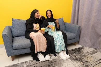 نشاط منزلي ترفيهي جماعي, المتعة في تناول الفشار اثناء مشاهدة التلفاز، قضاء اوقات ممتعة بين الاصدقاء, إمرأتان عربيتان خليجيتان اماراتيان ترتديان العباءه والحجاب تجلسان على الكنبة تشاهدان فيلم مسلي بايماءات الاندماج و السرور، الاستمتاع باوقات الفراغ