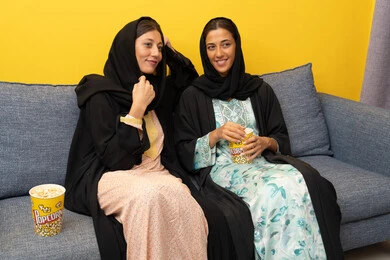 المتعة في تناول الفشار اثناء مشاهدة التلفاز، قضاء اوقات ممتعة بين الاصدقاء, إمرأتان عربيتان خليجيتان اماراتيان ترتديان العباءه والحجاب تجلسان على الكنبة تشاهدان فيلم مسلي بايماءات الاندماج و السرور، الاستمتاع باوقات الفراغ، نشاط منزلي ترفيهي جماعي