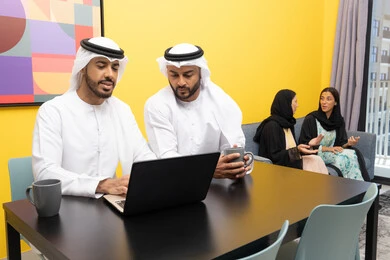 Two women wearing abaya and hijab exchanging conversations, managing and organizing business affairs via laptop, enjoying sitting at home, using a modern and advanced technical device, two Arab Gulf Emirati men wearing kandura and ghutra working on laptops, completing work tasks remotely