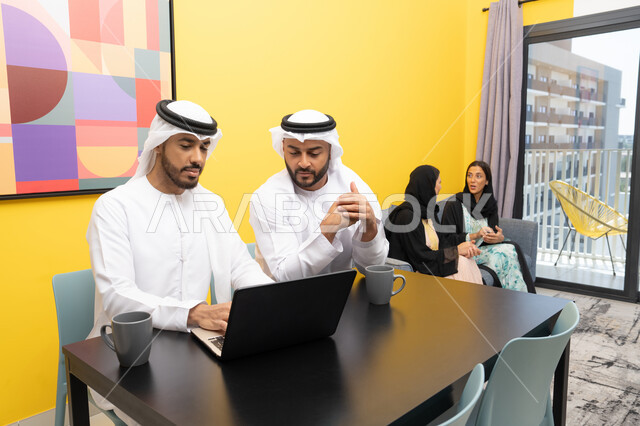 إنجاز مهام العمل عن بعد، إمرأتان ترتديان العباءة والحجاب تتبادلان الاحاديث، إدارة شؤون الأعمال وتنظيمها عبر اللابتوب، الاستمتاع بالجلوس في المنزل، استخدام جهاز تقني حديث ومتطور، رجلان عربيان خليجيان اماراتيان يرتديان الكندورة والغترة يعملان على الحاسوب المحمول