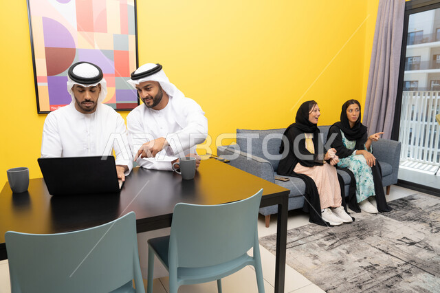 إمرأتان ترتديان العباءة والحجاب تتبادلان الاحاديث، إدارة شؤون الأعمال وتنظيمها عبر اللابتوب، الاستمتاع بالجلوس في المنزل، استخدام جهاز تقني حديث ومتطور، رجلان عربيان خليجيان اماراتيان يرتديان الكندورة والغترة يعملان على الحاسوب المحمول، إنجاز مهام العمل عن بعد