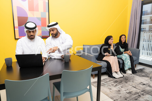 الاستمتاع بالجلوس في المنزل، استخدام جهاز تقني حديث ومتطور، رجلان عربيان خليجيان اماراتيان يرتديان الكندورة والغترة يعملان على الحاسوب المحمول، إنجاز مهام العمل عن بعد، إمرأتان ترتديان العباءة والحجاب تتبادلان الاحاديث، إدارة شؤون الأعمال وتنظيمها عبر اللابتوب