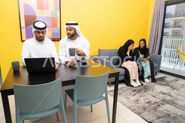 اتمام مهام العمل عن بعد، إمرأتان ترتديان العباءة والحجاب تتبادلان الاحاديث، إدارة شؤون الأعمال وتنظيمها عبر اللابتوب، الاستمتاع بالجلوس في المنزل، استخدام جهاز تقني حديث ومتطور، رجلان عربيان خليجيان اماراتيان يرتديان الكندورة والغترة يعملان على الحاسوب المحمول