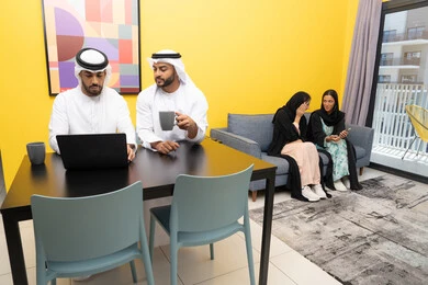 Completing work tasks remotely, two women wearing abaya and hijab exchanging conversations, managing and organizing business affairs via laptop, enjoying sitting at home, using a modern and advanced technical device, two Emirati Gulf Arab men wearing kandura and ghutra working on laptops
