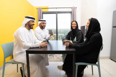 Enjoying hot drinks, a quiet family gathering at home, two Emirati Gulf Arab men wearing kanduras and ghutras sitting at the table with their wives with expressions of pleasure and happiness, exchanging news and interesting conversations, the concept of harmony and entertainment