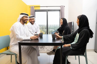 جلسة عائلية هادئة في المنزل، رجلان عربيان خليجيان اماراتيان يرتديان الكندورة والغترة يجلسان على الطاولة مع زوجاتهم بتعابير السرور والسعادة، تبادل الاخبار والاحاديث الممتعة، مفهوم الانسجام والتسلية، الاستمتاع بتناول المشروبات الساخنة