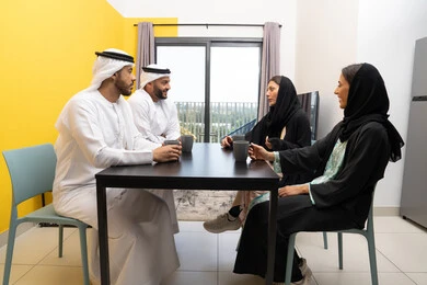 مفهوم الانسجام والتسلية، الاستمتاع بتناول المشروبات الساخنة ، جلسة عائلية هادئة في المنزل، رجلان عربيان خليجيان اماراتيان يرتديان الكندورة والغترة يجلسان على الطاولة مع زوجاتهم بتعابير السرور والسعادة، تبادل الاخبار والاحاديث الممتعة