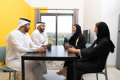 الاستمتاع بتناول المشروبات الساخنة ، جلسة عائلية هادئة في المنزل، رجلان عربيان خليجيان اماراتيان يرتديان الكندورة والغترة يجلسان على الطاولة مع زوجاتهم بتعابير السرور والسعادة، تبادل الاخبار والاحاديث الممتعة، مفهوم الانسجام والتسلية