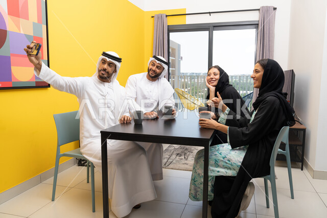 تطوير العلاقات الإجتماعية، ايماءات السعادة والسرور، قضاء وقت ممتع برفقة الأصدقاء، التقاط صور التذكارية عبر الجوال، اصدقاء عرب خليجيون اماراتيون يرتدون الزي التقليدي يقومون بالتجمع على الطاولة فى المنزل، مفهوم التقنيات والأجهزة الحديثة
