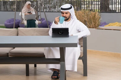الاستمتاع بالمشروبات الساخنه ،مفهوم التكنولوجيا وإستخدام تقنيات حديثة ، استخدم جهاز التابلت لانجاز المشاريع والمهام ، اجراء مكالمه فيديو،  قضاء وقت ممتع فى الخارج ،شاب عربي خليجي إماراتي يرتدي الكندورة والغترة البيضاء يجلس في المقهى ، تجلس فى الخلف سيدة اماراتية تستخدم اللابتوب