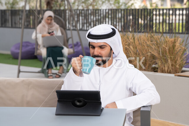 مفهوم التكنولوجيا وإستخدام تقنيات حديثة ، استخدم جهاز التابلت لانجاز المشاريع والمهام ، اجراء مكالمه فيديو،  قضاء وقت ممتع فى الخارج ،شاب عربي خليجي إماراتي يرتدي الكندورة والغترة البيضاء يجلس في المقهى ، تجلس فى الخلف سيدة اماراتية تستخدم اللابتوب ، الاستمتاع بالمشروبات الساخنه