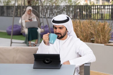 مفهوم التكنولوجيا وإستخدام تقنيات حديثة ، استخدم جهاز التابلت لانجاز المشاريع والمهام ، اجراء مكالمه فيديو،  قضاء وقت ممتع فى الخارج ،شاب عربي خليجي إماراتي يرتدي الكندورة والغترة البيضاء يجلس في المقهى ، تجلس فى الخلف سيدة اماراتية تستخدم اللابتوب ، الاستمتاع بالمشروبات الساخنه
