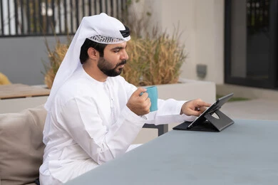شاب عربي خليجي إماراتي يرتدي الكندورة والغترة البيضاء يجلس في المقهى ، قضاء وقت ممتع فى الخارج ،مفهوم التكنولوجيا وإستخدام تقنيات حديثة،الاستمتاع بالمشروبات الساخنه ، اجراء مكالمه فيديو، استخدم جهاز التابلت لانجاز المشاريع والمهام