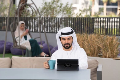 استخدم جهاز التابلت لانجاز المشاريع والمهام ، اجراء مكالمه فيديو،  قضاء وقت ممتع فى الخارج ،شاب عربي خليجي إماراتي يرتدي الكندورة والغترة البيضاء يجلس في المقهى ، تجلس فى الخلف سيدة اماراتية تستخدم الجوال، مفهوم التكنولوجيا وإستخدام تقنيات حديثة الاستمتاع بالمشروبات الساخنه