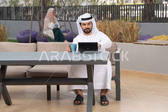 اجراء مكالمه فيديو،  قضاء وقت ممتع فى الخارج ،شاب عربي خليجي إماراتي يرتدي الكندورة والغترة البيضاء يجلس في المقهى ، تجلس فى الخلف سيدة اماراتية تستخدم الجوال، مفهوم التكنولوجيا وإستخدام تقنيات حديثة الاستمتاع بالمشروبات الساخنه ، استخدم جهاز التابلت لانجاز المشاريع والمهام