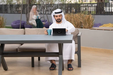 اجراء مكالمه فيديو،  قضاء وقت ممتع فى الخارج ،شاب عربي خليجي إماراتي يرتدي الكندورة والغترة البيضاء يجلس في المقهى ، تجلس فى الخلف سيدة اماراتية تستخدم الجوال، مفهوم التكنولوجيا وإستخدام تقنيات حديثة الاستمتاع بالمشروبات الساخنه ، استخدم جهاز التابلت لانجاز المشاريع والمهام