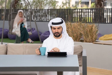 مفهوم التكنولوجيا وإستخدام تقنيات حديثة، الاستمتاع بالمشروبات الساخنه ، اجراء مكالمه فيديو، قضاء وقت ممتع فى الخارج ، استخدم جهاز التابلت لانجاز المشاريع والمهام ، شاب عربي خليجي إماراتي يرتدي الكندورة والغترة البيضاء يجلس في المقهى ، تجلس فى الخلف سيدة اماراتية تستخدم الجوال