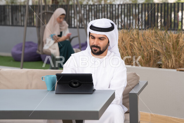 الاستمتاع بالمشروبات الساخنه ، اجراء مكالمه فيديو، قضاء وقت ممتع فى الخارج ، استخدم جهاز التابلت لانجاز المشاريع والمهام ، شاب عربي خليجي إماراتي يرتدي الكندورة والغترة البيضاء يجلس في المقهى ، تجلس فى الخلف سيدة اماراتية تستخدم الجوال، مفهوم التكنولوجيا وإستخدام تقنيات حديثة