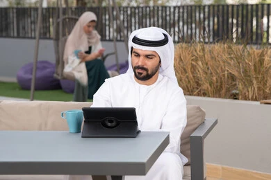 Enjoying hot drinks, making a video call, having a good time outside, using a tablet to complete projects and tasks, a young Emirati Gulf Arab man wearing a white kandura and ghutra sitting in a cafe, an Emirati woman sitting in the back using a mobile phone, the concept of technology and the use of modern techniques