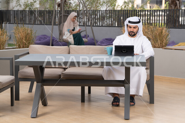Having fun outside, using a tablet to complete projects and tasks, a young Emirati Gulf Arab man wearing a white kandura and ghutra sitting in a cafe, an Emirati woman sitting in the back using a mobile phone, the concept of technology and the use of modern techniques, enjoying hot drinks, making a video call