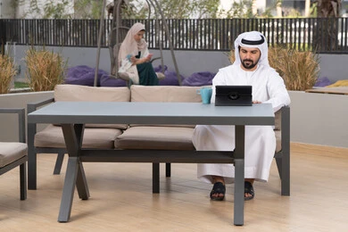 Having fun outside, using a tablet to complete projects and tasks, a young Emirati Gulf Arab man wearing a white kandura and ghutra sitting in a cafe, an Emirati woman sitting in the back using a mobile phone, the concept of technology and the use of modern techniques, enjoying hot drinks, making a video call