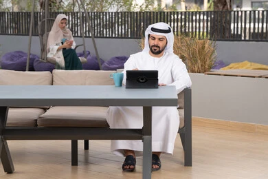 Using a tablet to complete projects and tasks, a young Emirati Gulf Arab man wearing a white kandura and ghutra sitting in a cafe, an Emirati woman sitting in the back using a mobile phone, spending a pleasant time outside, the concept of technology and the use of modern techniques, enjoying hot drinks, making a video call