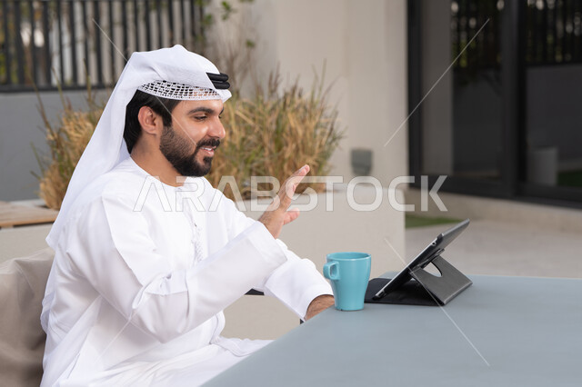 مفهوم التكنولوجيا وإستخدام تقنيات حديثة،الاستمتاع بالمشروبات الساخنه ، اجراء مكالمه فيديو، استخدم جهاز التابلت لانجاز المشاريع والمهام ،  قضاء وقت ممتع فى الخارج ،شاب عربي خليجي إماراتي يرتدي الكندورة والغترة البيضاء يجلس في المقهى