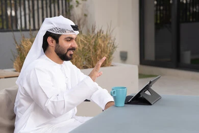 The concept of technology and the use of modern techniques, enjoying hot drinks, making a video call, using a tablet to complete projects and tasks, spending a pleasant time outside, an Emirati Gulf Arab young man wearing a white kandura and ghutra sitting in a cafe