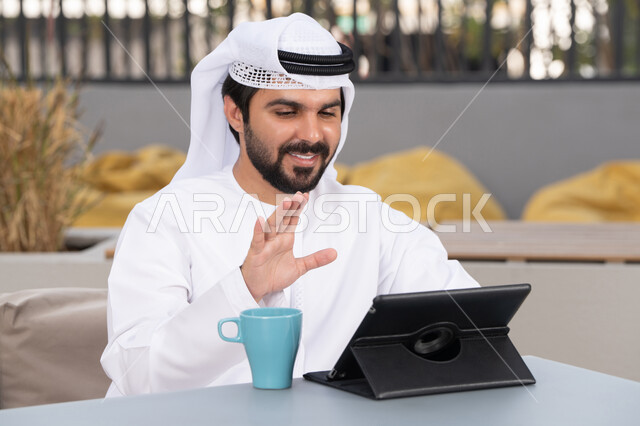 قضاء وقت ممتع فى الخارج ،مفهوم التكنولوجيا وإستخدام تقنيات حديثة،الاستمتاع بالمشروبات الساخنه ، اجراء مكالمه فيديو، استخدم جهاز التابلت لانجاز المشاريع والمهام ، شاب عربي خليجي إماراتي يرتدي الكندورة والغترة البيضاء يجلس في المقهى