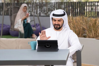 شاب عربي خليجي إماراتي يرتدي الكندورة والغترة البيضاء يجلس في المقهى ، تجلس فى الخلف سيدة اماراتية تستخدم الجوال، قضاء وقت ممتع فى الخارج ،مفهوم التكنولوجيا وإستخدام تقنيات حديثة،الاستمتاع بالمشروبات الساخنه ، اجراء مكالمه فيديو، استخدم جهاز التابلت لانجاز المشاريع والمهام