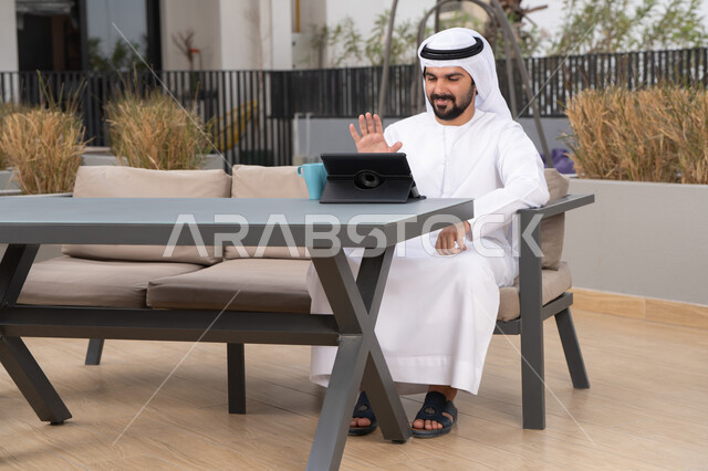 الاستمتاع بالمشروبات الساخنه ، اجراء مكالمه فيديو، استخدم جهاز التابلت لانجاز المشاريع والمهام ، مفهوم التكنولوجيا وإستخدام تقنيات حديثة، قضاء وقت ممتع فى الخارج ،شاب عربي خليجي إماراتي يرتدي الكندورة والغترة البيضاء يجلس في المقهى