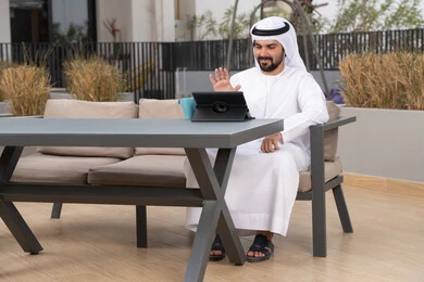 الاستمتاع بالمشروبات الساخنه ، اجراء مكالمه فيديو، استخدم جهاز التابلت لانجاز المشاريع والمهام ، مفهوم التكنولوجيا وإستخدام تقنيات حديثة، قضاء وقت ممتع فى الخارج ،شاب عربي خليجي إماراتي يرتدي الكندورة والغترة البيضاء يجلس في المقهى