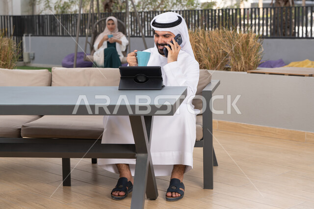 اجراء مكالمه هاتفيه، قضاء وقت ممتع فى الخارج ، الاستمتاع بالمشروبات الساخنه ، شاب عربي خليجي إماراتي يرتدي الكندورة والغترة البيضاء يجلس في المقهى ، تجلس فى الخلف سيدة اماراتيه تستخدم الجوال ، استخدم جهاز الحاسوب المحمول لانجاز المشاريع والمهام ، مفهوم التكنولوجيا وإستخدام تقنيات حديثة