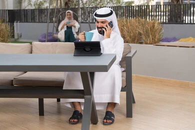 Making a phone call, having a good time outside, enjoying a hot drink, a young Emirati Gulf Arab man wearing a white kandura and ghutra sitting in a cafe, an Emirati woman sitting in the back using a mobile phone, using a laptop to complete projects and tasks, the concept of technology and the use of modern techniques