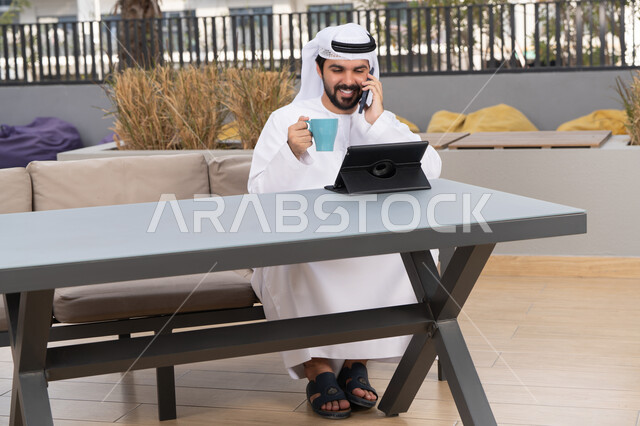 استخدم جهاز الحاسوب المحمول لانجاز المشاريع والمهام ،الاستمتاع بالمشروبات الساخنه ، مفهوم التكنولوجيا وإستخدام تقنيات حديثة، اجراء مكالمه هاتفيه، قضاء وقت ممتع فى الخارج ،شاب عربي خليجي إماراتي يرتدي الكندورة والغترة البيضاء يجلس في المقهى