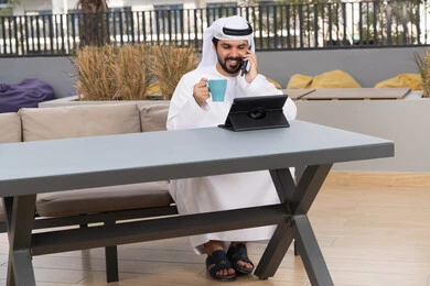 Use laptop to complete projects and tasks, enjoy hot drinks, concept of technology and use of modern techniques, make a phone call, spend a pleasant time outside, young Arab Gulf Emirati man wearing white kandura and ghutra sitting in a cafe