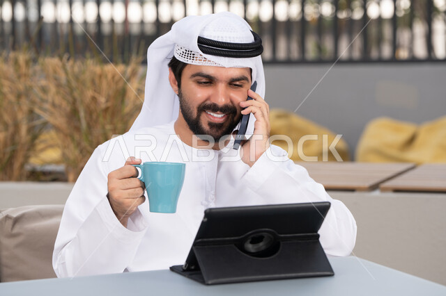 الاستمتاع بالمشروبات الساخنه ، مفهوم التكنولوجيا وإستخدام تقنيات حديثة، اجراء مكالمه هاتفيه، قضاء وقت ممتع فى الخارج ،شاب عربي خليجي إماراتي يرتدي الكندورة والغترة البيضاء يجلس في المقهى ، استخدم جهاز الحاسوب المحمول لانجاز المشاريع والمهام