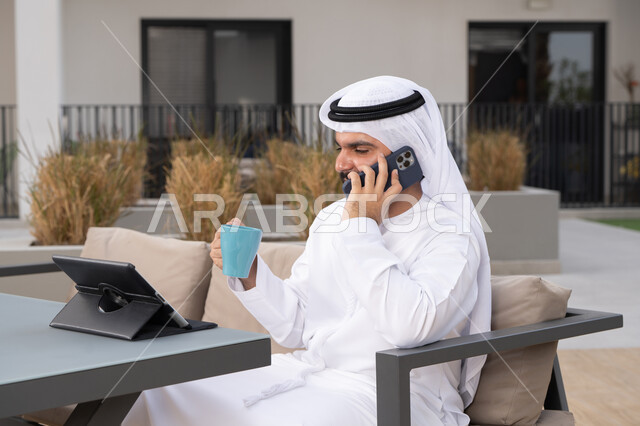 مفهوم التكنولوجيا وإستخدام تقنيات حديثة، اجراء مكالمه هاتفيه، قضاء وقت ممتع فى الخارج ، الاستمتاع بالمشروبات الساخنه ، شاب عربي خليجي إماراتي يرتدي الكندورة والغترة البيضاء يجلس في المقهى ، استخدم جهاز الحاسوب المحمول لانجاز المشاريع والمهام