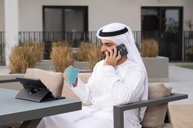 مفهوم التكنولوجيا وإستخدام تقنيات حديثة، اجراء مكالمه هاتفيه، قضاء وقت ممتع فى الخارج ، الاستمتاع بالمشروبات الساخنه ، شاب عربي خليجي إماراتي يرتدي الكندورة والغترة البيضاء يجلس في المقهى ، استخدم جهاز الحاسوب المحمول لانجاز المشاريع والمهام