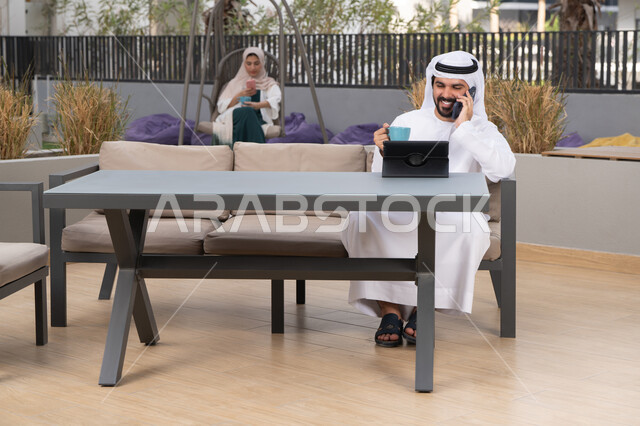 قضاء وقت ممتع فى الخارج ، الاستمتاع بالمشروبات الساخنه ، كوفي شوب راقي ، شاب عربي خليجي إماراتي يرتدي الكندورة والغترة البيضاء يجلس في المقهى ، تجلس فى الخلف سيدة اماراتيه تستخدم الجوال ، استخدم جهاز الحاسوب المحمول لانجاز المشاريع والمهام ، مفهوم التكنولوجيا وإستخدام تقنيات حديثة