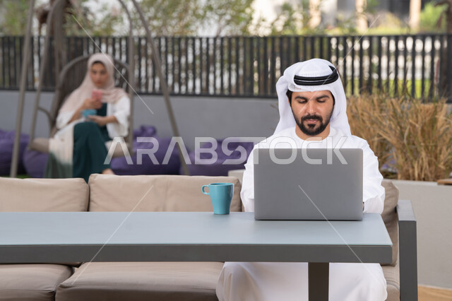 الاستمتاع بالمشروبات الساخنه ، كوفي شوب راقي ، شاب عربي خليجي إماراتي يرتدي الكندورة والغترة البيضاء يجلس في المقهى ، تجلس فى الخلف سيدة اماراتيه تستخدم الجوال ، استخدم جهاز الحاسوب المحمول لانجاز المشاريع والمهام ، مفهوم التكنولوجيا وإستخدام تقنيات حديثة ، قضاء وقت ممتع فى الخارج