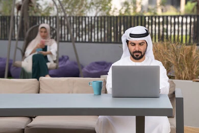Enjoying hot drinks, a classy coffee shop, a young Emirati Gulf Arab man wearing a white kandura and ghutra sitting in the cafe, an Emirati woman sitting in the back using a mobile phone, using a laptop to complete projects and tasks, the concept of technology and the use of modern techniques, spending a pleasant time outside