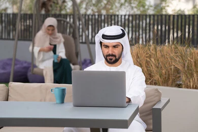 شاب عربي خليجي إماراتي يرتدي الكندورة والغترة البيضاء يجلس في المقهى ، تجلس فى الخلف سيدة اماراتيه تستخدم الجوال ، استخدم جهاز الحاسوب المحمول لانجاز المشاريع والمهام في الكافيه ، مفهوم التكنولوجيا وإستخدام تقنيات حديثة ، قضاء وقت ممتع فى الخارج ، الاستمتاع بالمشروبات الساخنه