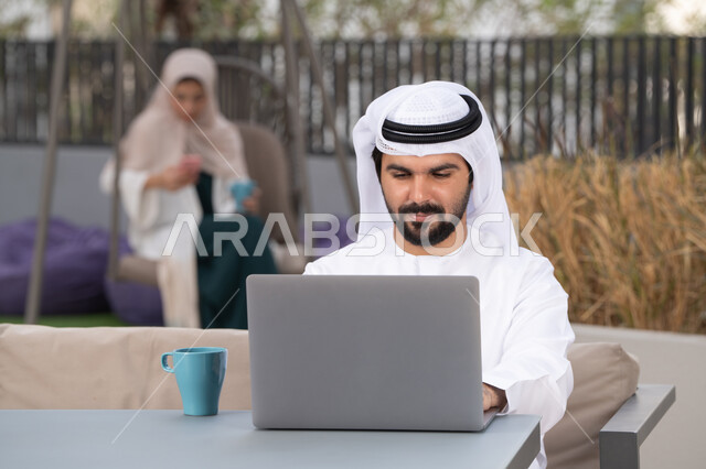 Using a laptop to complete projects and tasks in a cafe, the concept of technology and the use of modern techniques, spending a pleasant time outside, enjoying hot drinks, a young Emirati Gulf Arab man wearing a white kandura and ghutra sitting in a cafe, an Emirati woman sitting in the back using a mobile phone
