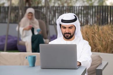 استخدم جهاز الحاسوب المحمول لانجاز المشاريع والمهام في الكافيه ، مفهوم التكنولوجيا وإستخدام تقنيات حديثة ، قضاء وقت ممتع فى الخارج ، الاستمتاع بالمشروبات الساخنه ، شاب عربي خليجي إماراتي يرتدي الكندورة والغترة البيضاء يجلس في المقهى ، تجلس فى الخلف سيدة اماراتيه تستخدم الجوال