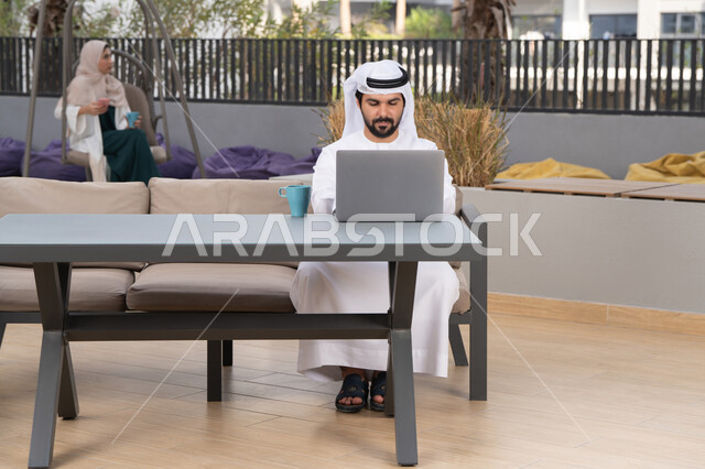 مفهوم التكنولوجيا وإستخدام تقنيات حديثة ، قضاء وقت ممتع فى الخارج ، الاستمتاع بالمشروبات الساخنه ،استخدم جهاز الحاسوب المحمول لانجاز المشاريع والمهام في الكافيه ، شاب عربي خليجي إماراتي يرتدي الكندورة والغترة البيضاء يجلس في المقهى ، تجلس فى الخلف سيدة اماراتيه تجري مكالمه هاتفيه