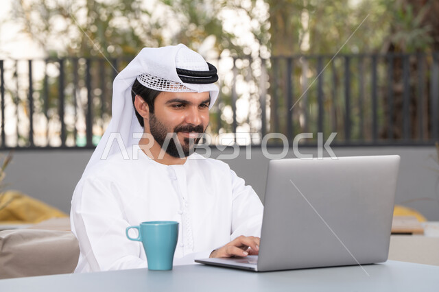 قضاء وقت ممتع فى الخارج ، الاستمتاع بالمشروبات الساخنه ، شاب عربي خليجي إماراتي يرتدي الكندورة والغترة البيضاء يجلس في المقهى ، استخدم جهاز الحاسوب المحمول لانجاز المشاريع والمهام ، مفهوم التكنولوجيا وإستخدام تقنيات حديثة
