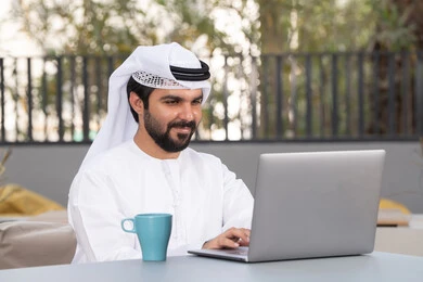 Spending a pleasant time outside, enjoying a hot beverage, a young Emirati Gulf Arab man wearing a white kandura and ghutra sitting in a café, using a laptop to complete projects and tasks, the concept of technology and the use of modern techniques.